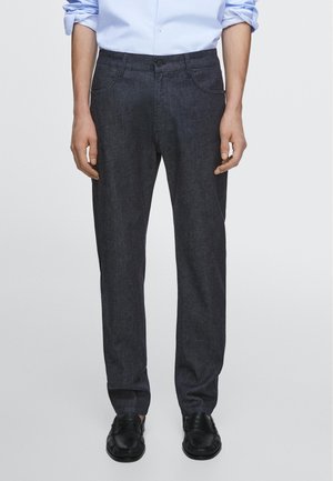 Man draagt donkergrijze broek met rechte pijpen, lichtblauw opgerolde overhemd en zwarte loafers, staand tegen een effen achtergrond.