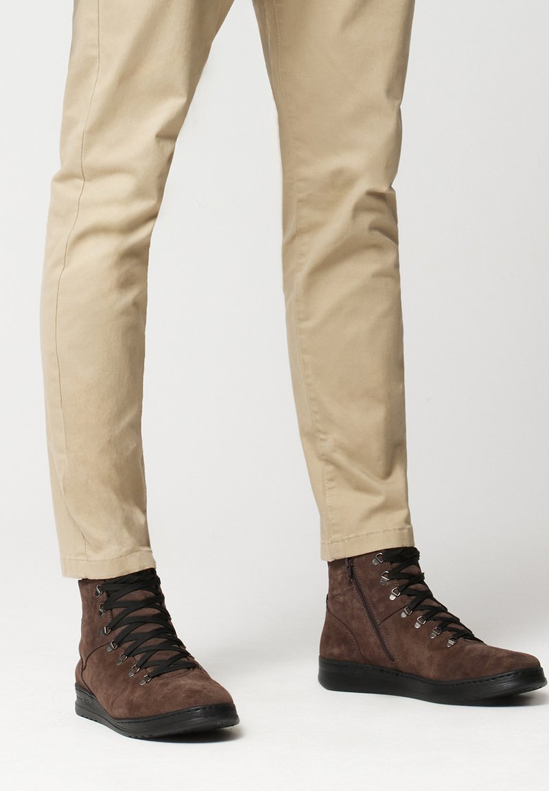 Brązowe zamszowe botki za kostkę z czarnymi sznurowadłami i metalowymi oczkami, zestawione z jasnobeżowymi spodniami w wersji tapered. Prosty design z teksturowanym wykończeniem.