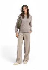 Hellbrauner Pullover mit Muster, beige weit geschnittene Hosen und helle Schuhe. Hält eine olivgrüne Umhängetasche. Weiche Textur, lockere Passform.