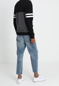 Personne portant un t-shirt à manches longues noir et gris avec des rayures blanches, un jean bleu clair et des baskets blanches Reebok, debout de dos.