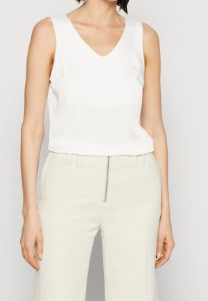 Femme portant un haut blanc sans manches à col en V et un pantalon blanc taille haute avec une fermeture éclair devant, sur un fond uni.