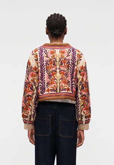 Femme aux cheveux nattés portant une veste courte beige ornée de broderies florales colorées et un pantalon en jean foncé, vue de dos.