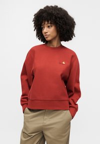 Γυναίκα με σγουρά μαλλιά, φορώντας κόκκινο φούτερ Carhartt και μπεζ παντελόνι, στέκεται με τα χέρια στις τσέπες, κοιτώντας προς το πλάι.