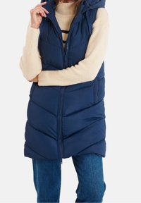 Gilet invernale blu navy con design trapuntato, cappuccio e zip frontale, indossato sopra un maglione beige, abbinato a jeans blu.