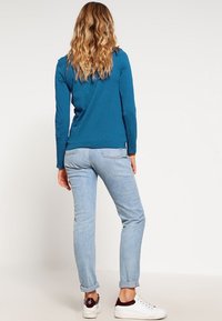Teal långärmad tröja i kombination med ljusblå jeans. Outfiten har en avslappnad passform och enkla vita sneakers med burgundyröda accenter.