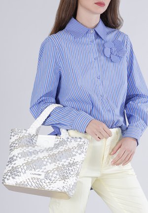 Mujer con camisa de botones a rayas azul y blanca con detalle de flores, sosteniendo un bolso blanco tejido, vestida con pantalones crema.