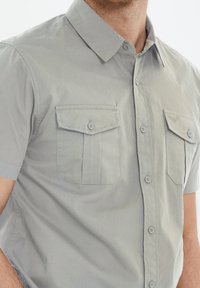 Camisa de manga corta gris claro hecha de tejido suave, con dos bolsillos en el pecho con solapa y cierre de botón, y un cuello tipo polo.