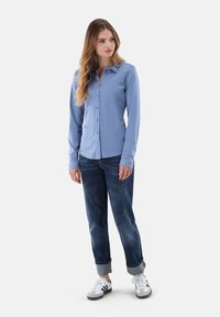 Helles blaues, langärmliges Hemd, kombiniert mit dunkelblauen Jeans und weißen Sneakers, stehend vor einem schlichten Hintergrund.