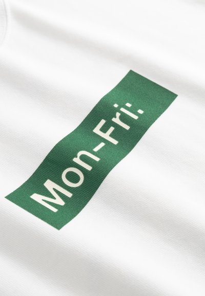 Hvit bomulls-t-skjorte med et rektangulært grønt stoffmerke med teksten «Man-Fre:» i hvitt, plassert sentralt på stoffet.