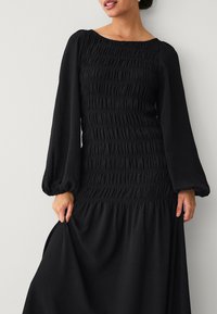 Schwarzes Kleid mit einem gesmockschnittenen Oberteil und langen, puffigen Ärmeln. Volle, fließende Röcke und strukturierter Stoff. Schlichtes, elegantes Design ohne Muster.