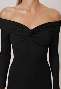 Top nero scollato sulle spalle con maniche lunghe, caratterizzato da un dettaglio annodato al centro e una texture a coste. Design liscio e aderente.