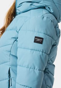 Veste matelassée bleu clair avec capuche, écusson logo Swedemount noir sur le haut du bras, portée par une personne aux longs cheveux châtains clairs.