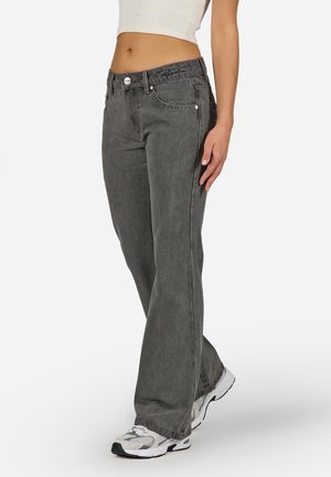 Person trägt locker sitzende ausgewaschene schwarze Jeans, weißes kurzes Tanktop und weiße Sportschuhe mit grauen und schwarzen Details, steht auf weißem Hintergrund.