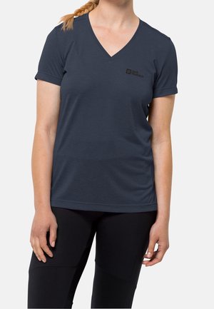 Kobieta ubrana w granatowy T-shirt z krótkim rękawem i dekoltem w serek oraz czarne, dopasowane spodnie, z włosami związanymi w boczny warkocz na jednolitym tle.