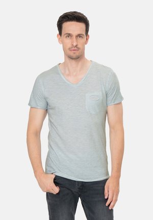 Uomo con capelli scuri che indossa una maglietta grigio chiaro con scollo a V e jeans scuri, in piedi con una mano in tasca davanti a uno sfondo neutro.