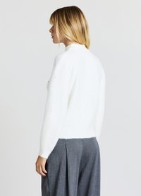 Pull blanc, doux, avec un col montant et des manches longues, orné d'accents en perles décoratives sur les manches. Associé à un pantalon plissé gris.