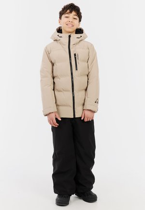 Beige gesteppte Jacke mit Kapuze und schwarzen Reißverschlüssen, mit einem Logo auf der Brust, kombiniert mit schwarzen Hosen und robusten schwarzen Stiefeln.