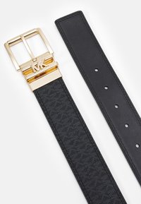 Zwarte leren riem met een gestructureerd oppervlak, voorzien van een gouden metalen gesp met de initialen "MK" en gaatjes voor aanpassing.