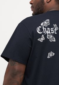 Svart t-skjorte med korte ermer, prydet med hvite broderte sommerfugler og teksten "Chase" på ryggen. Mykt bomullsmateriale.