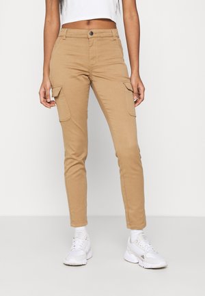 Kobieta w beżowych spodniach typu cargo z klapkowymi kieszeniami, białym krótkim topie, białych skarpetkach i białych trampkach, stojąca na jednolitym tle.