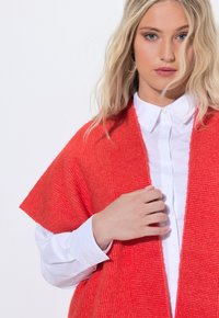 Röd stickad cardigan med vida ärmar, öppen front, strukturerad tyg, lager på lager över en vit skjorta med krage.