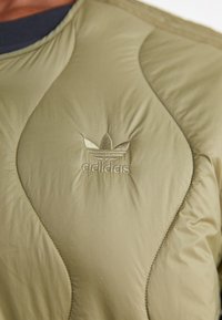 Svetlo olivno zelena jakna s kviltanim vzorcem in teksturiranimi valovitimi vzorci; vsebuje izšito Adidasovo logo v usklajeni barvi na levi strani.
