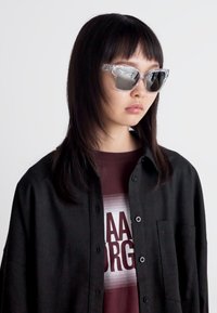 Ochelari de soare transparenti cu un cadru clar, având un model texturat ușor, purtați cu o cămașă neagră cu nasturi peste un tricou grafic în nuanță de vișinie.