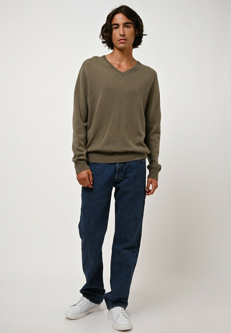 Khaki v-hals trui gemaakt van zacht materiaal, gecombineerd met losse blauwe denim jeans en witte sneakers, staande op een effen achtergrond.