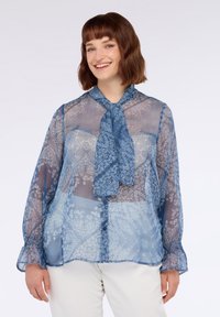 Blusa blu trasparente con motivo in pizzo floreale, dal davanti con bottoni e collo annodato. Maniche lunghe con polsini a volant, texture leggera e fluttuante.