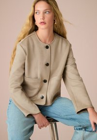 Veste beige courte avec un col rond, des poches avant et des boutons noirs. Modèle portant un jean bleu clair, assis sur un tabouret.