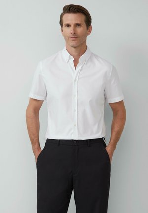 Chemise blanche à manches courtes et boutonnière avec un col, en tissu lisse, associée à un pantalon noir, présentant une coupe classique.