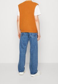 Orange gestreifter ärmelloser Strickpullover über einem weißen T-Shirt, kombiniert mit blauen weit geschnittenen Jeans und weißen Sneakern.