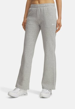 Person trägt graue Jogginghose mit weißen Seitenstreifen und weißen Sportschuhen, steht vor einem schlichten hellen Hintergrund.