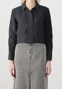 Czarna krótka kurtka zapinana z przodu na guziki, z dwoma kieszeniami na piersi i długimi rękawami, w zestawie z jasnoszarą spódnicą dżinsową z przednim rozcięciem.