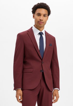 Man draagt een bordeaux pak met een wit overhemd en een donkerblauwe stropdas, staand tegen een effen witte achtergrond.