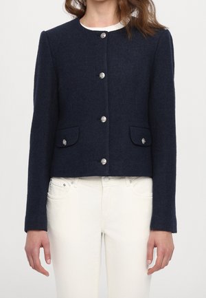 Kobieta ubrana w dopasowaną granatową wełnianą marynarkę z srebrnymi guzikami i klapowanymi kieszeniami, nosząca białe spodnie.