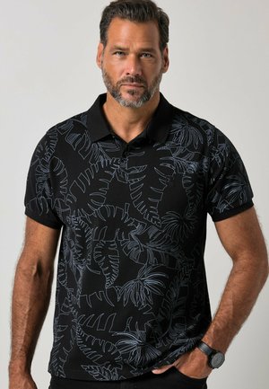 Mand med skæg iført sort poloshirt med lyse bladmønstre, stående med den ene hånd i lommen mod en ensfarvet baggrund.