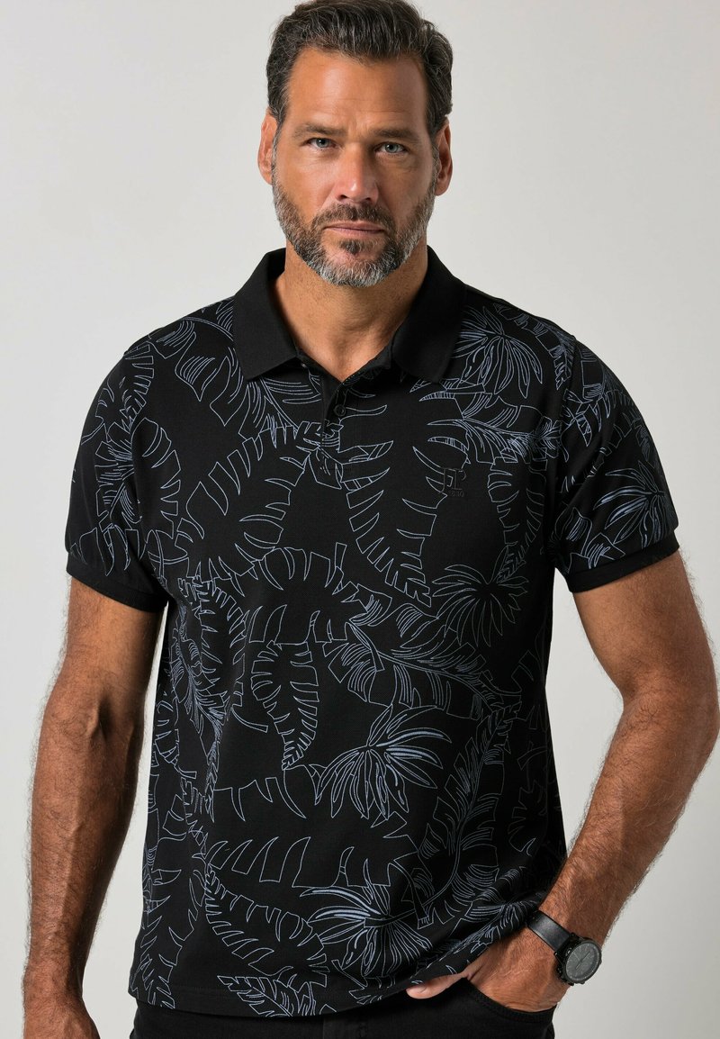 Man with beard wearing black polo shirt with light leaf patterns, standing with one hand in pocket against plain background.