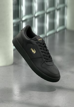 Black Lacoste sneaker with gold logo and laces, floating above a gray concrete floor with glass block wall background.