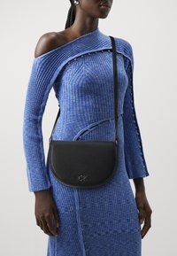 Czarna skórzana torebka typu crossbody o zaokrąglonym kształcie, z wytłoczonym logo "CK", noszona na teksturowanej niebieskiej sukience z prążkami i akcentami guzików.