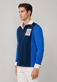Man wearing a blue and navy rugby shirt with white collared cuffs and white pants, standing against a plain background, facing left.