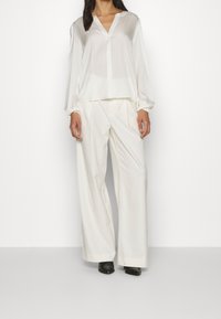 Weiße, leichte Bluse mit V-Ausschnitt und langen Ärmeln, kombiniert mit lockeren weißen Hosen und schwarzen Schuhen. Glatte Textur, minimalistisches Design.