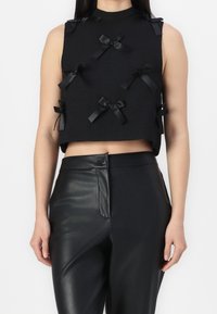 Top corto negro con un cuello alto, tejido texturizado y múltiples lazos de satén en la parte delantera. Se combina con pantalones de cuero negro de talle alto.
