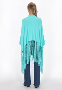 Pikkade juustega naine, kes kannab türkiissinist frantsiä�äridega poncho't, siniseid teksaseid ja musti kingi, seisab valge tausta against, seljaga vaataja poole.