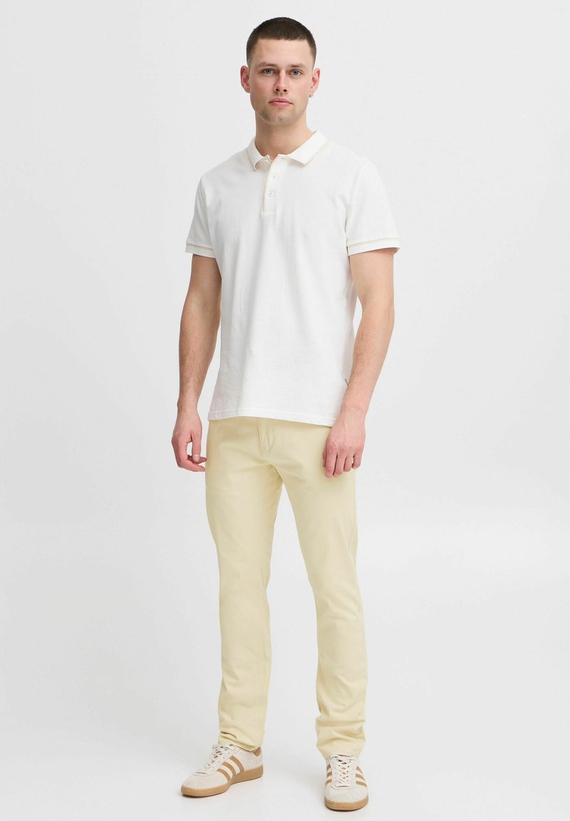Hvit polo skjorte med korte ermer og lysebeige detaljer, parret med lysegule bukser og beige joggesko med brune striper.