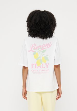 Άτομο με σγουρά μαλλιά που φοράει λευκό T-shirt με ροζ κείμενο "Limoni Italy Costa d'Amalfi" και γραφικό με κλαδί λεμονιάς στην πλάτη.