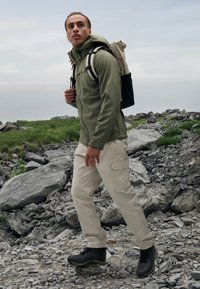 Zielona bluza z zamkiem i kapturem, beżowe spodnie cargo oraz czarne buty turystyczne. Osoba niesie jasny plecak. Teksturowana, skalista nawierzchnia.