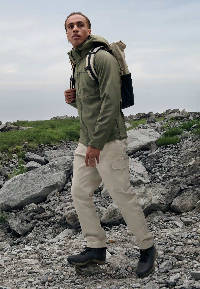 Chaqueta verde con cremallera y capucha, pantalones cargo beige y zapatos de senderismo negros. La persona lleva una mochila de color claro. Suelo rocoso y texturizado.