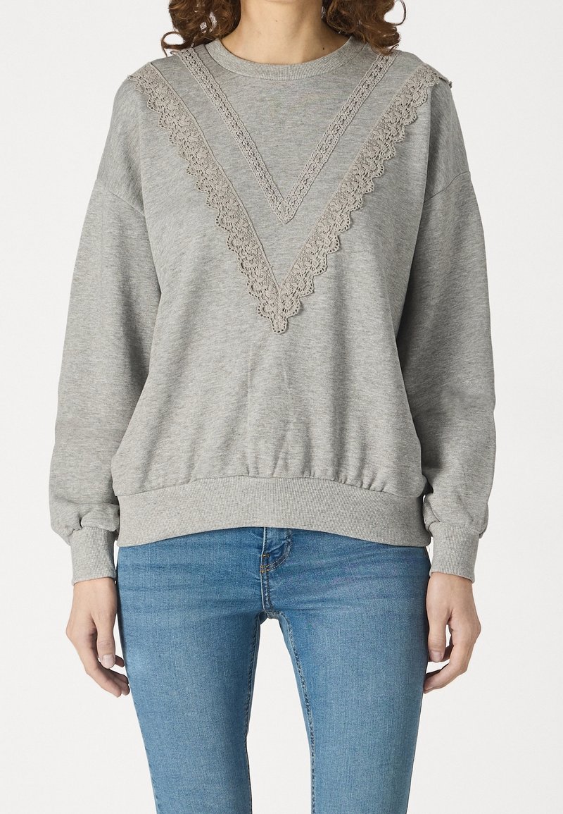 Femme portant un sweat-shirt gris clair avec une bordure en dentelle en forme de V sur la poitrine, associé à un jean bleu, debout devant un fond blanc.