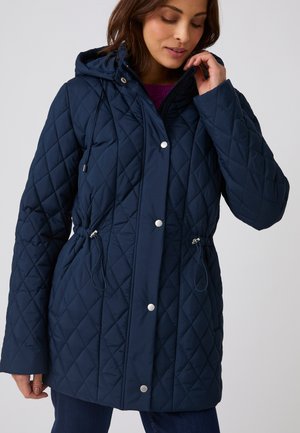 Femme portant une veste matelassée bleu marine avec capuche, taille cintrée par un cordon et boutons-pression, ajustant le col d’une main.